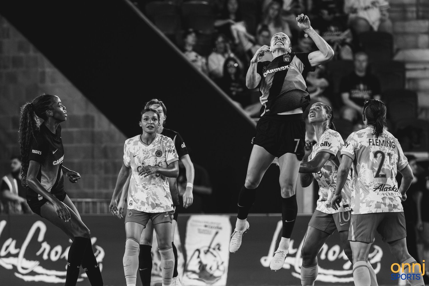 Black and white photo of female handball players competing in an intense match, with one player jumping high to catch the ball.