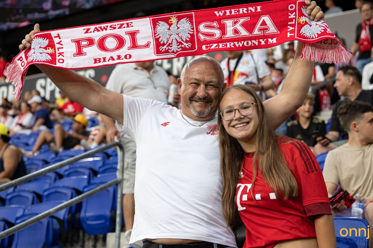 Two Polish football fans holding a red and white POLSKA scarf at a stadium, smiling and cheering for their team.