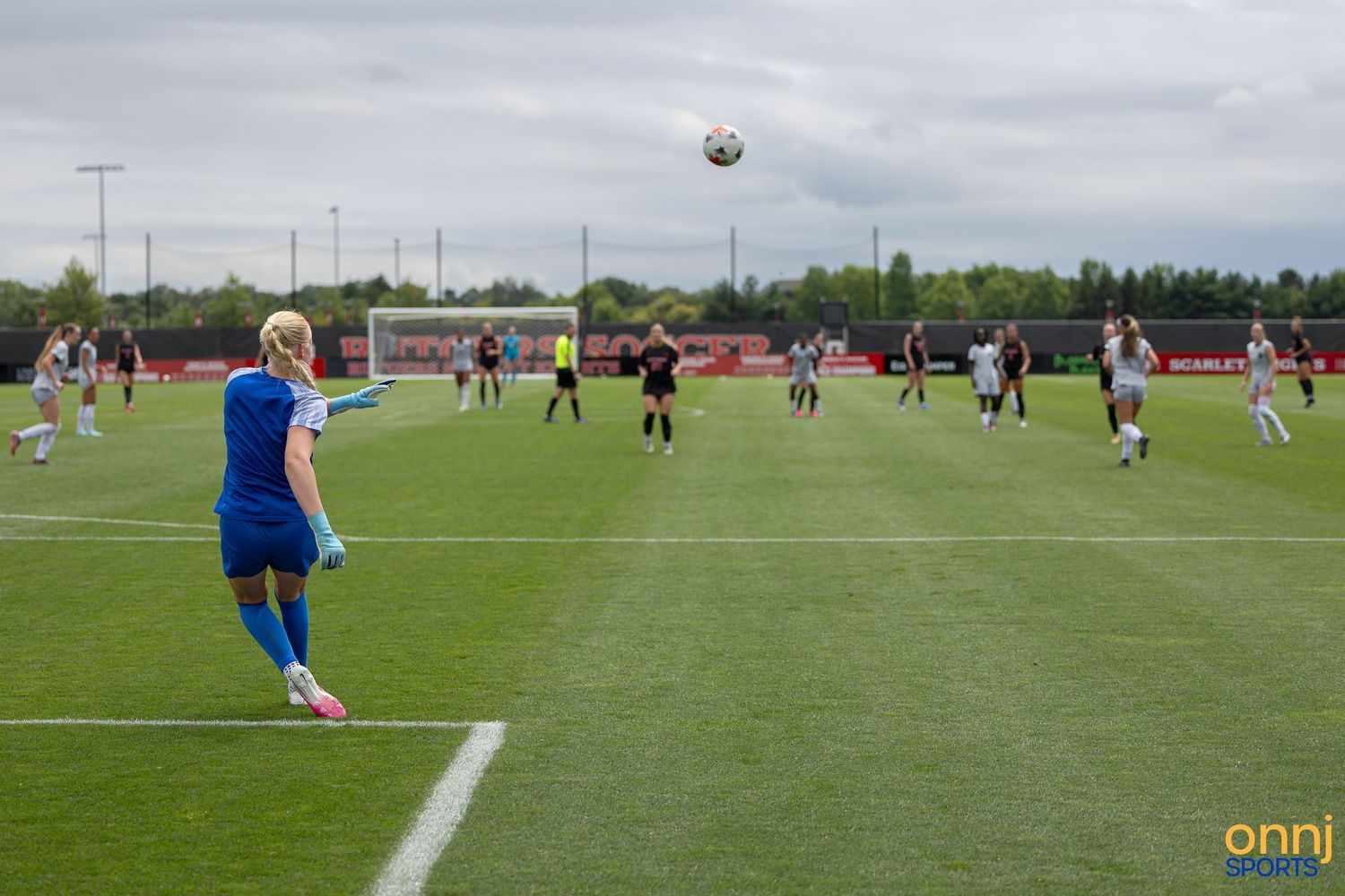 Female goalkeeper in blue kit kicks ball during women's soccer match on green pitch with players in background.