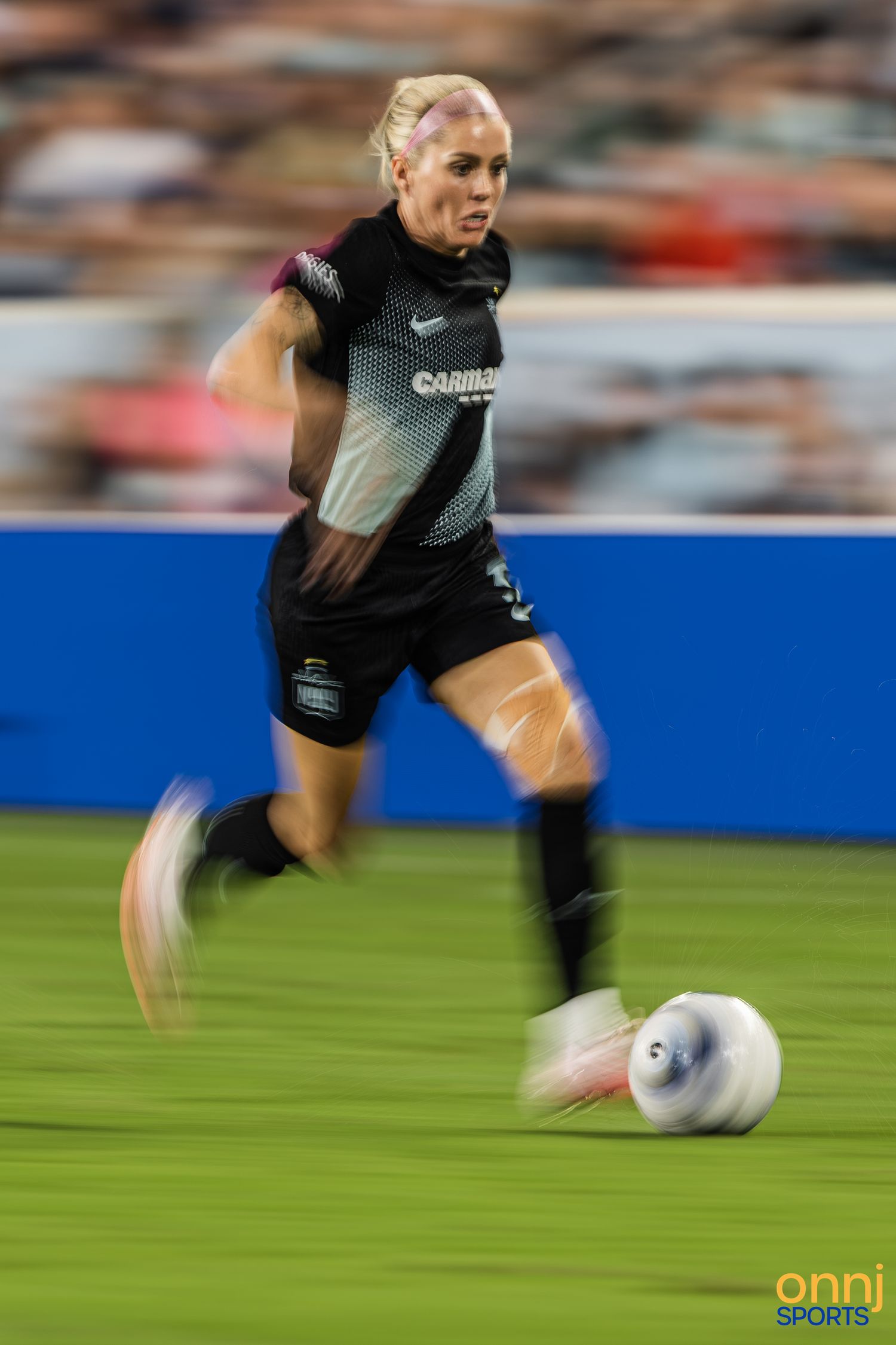 Female soccer player in black Nike kit dribbles ball at speed during a night match, motion blur effect.