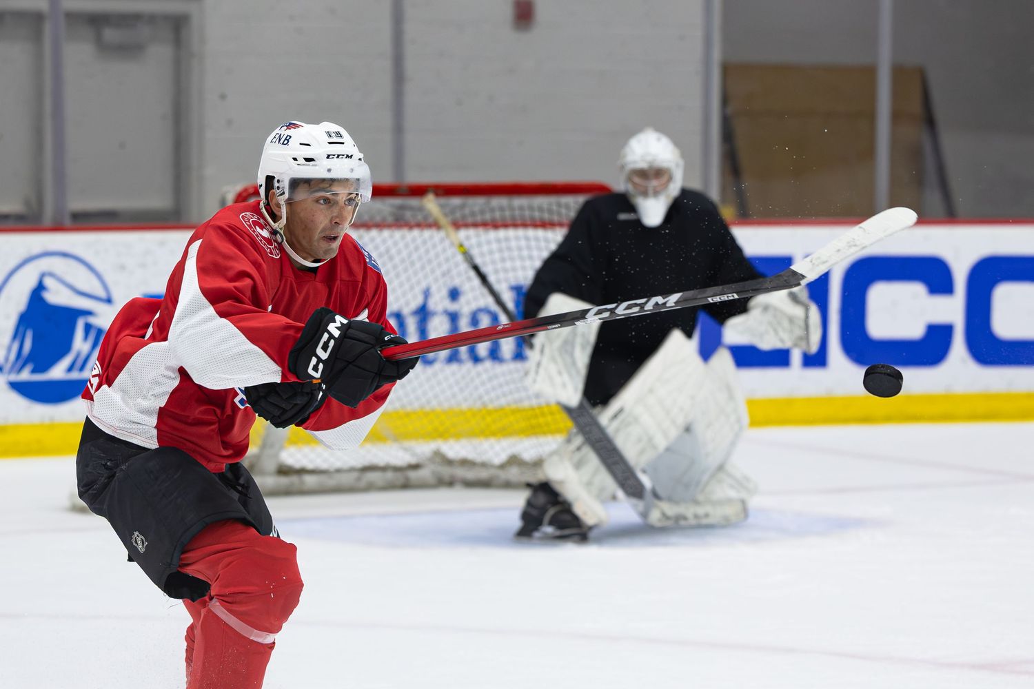 Hockey player in red jersey takes a shot on goal during an indoor ice rink practice session with goalie in background.