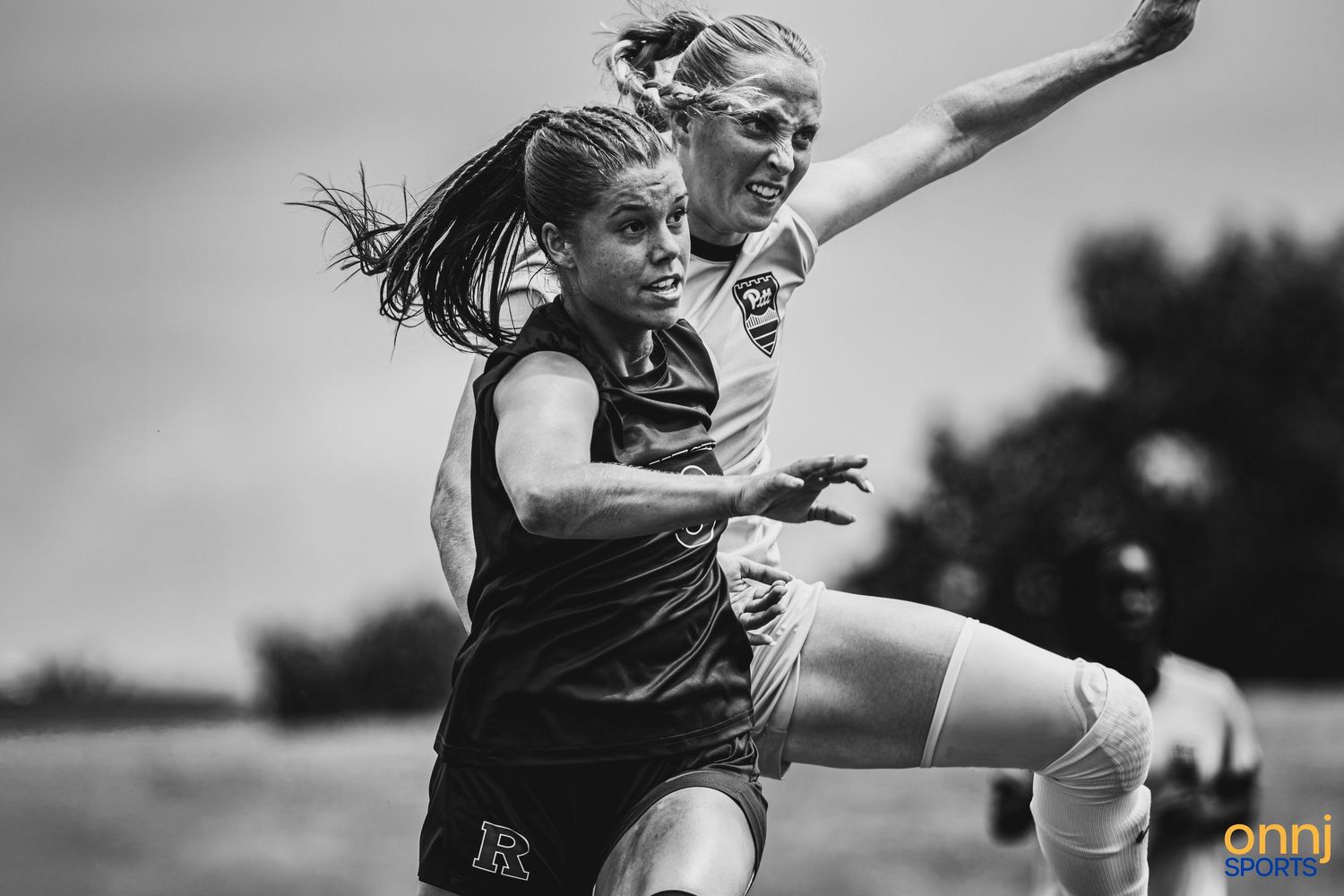 Two female soccer players compete in a black and white action photo during an outdoor match.