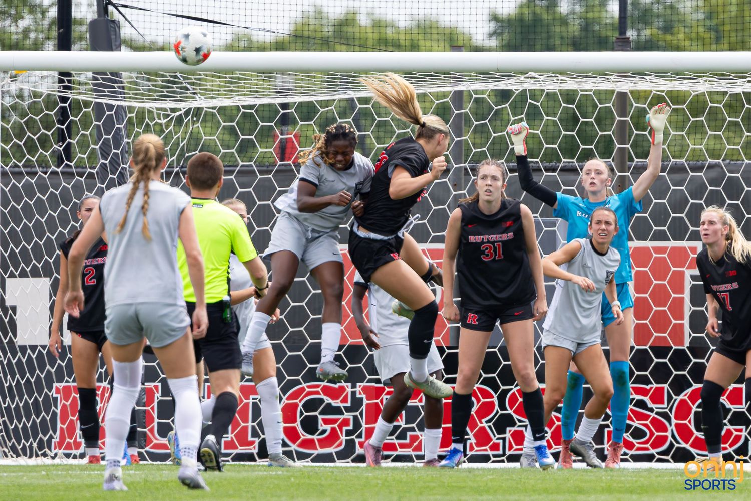 Women's college soccer players jump for a header near the goal during a Rutgers University match on a sunny day.