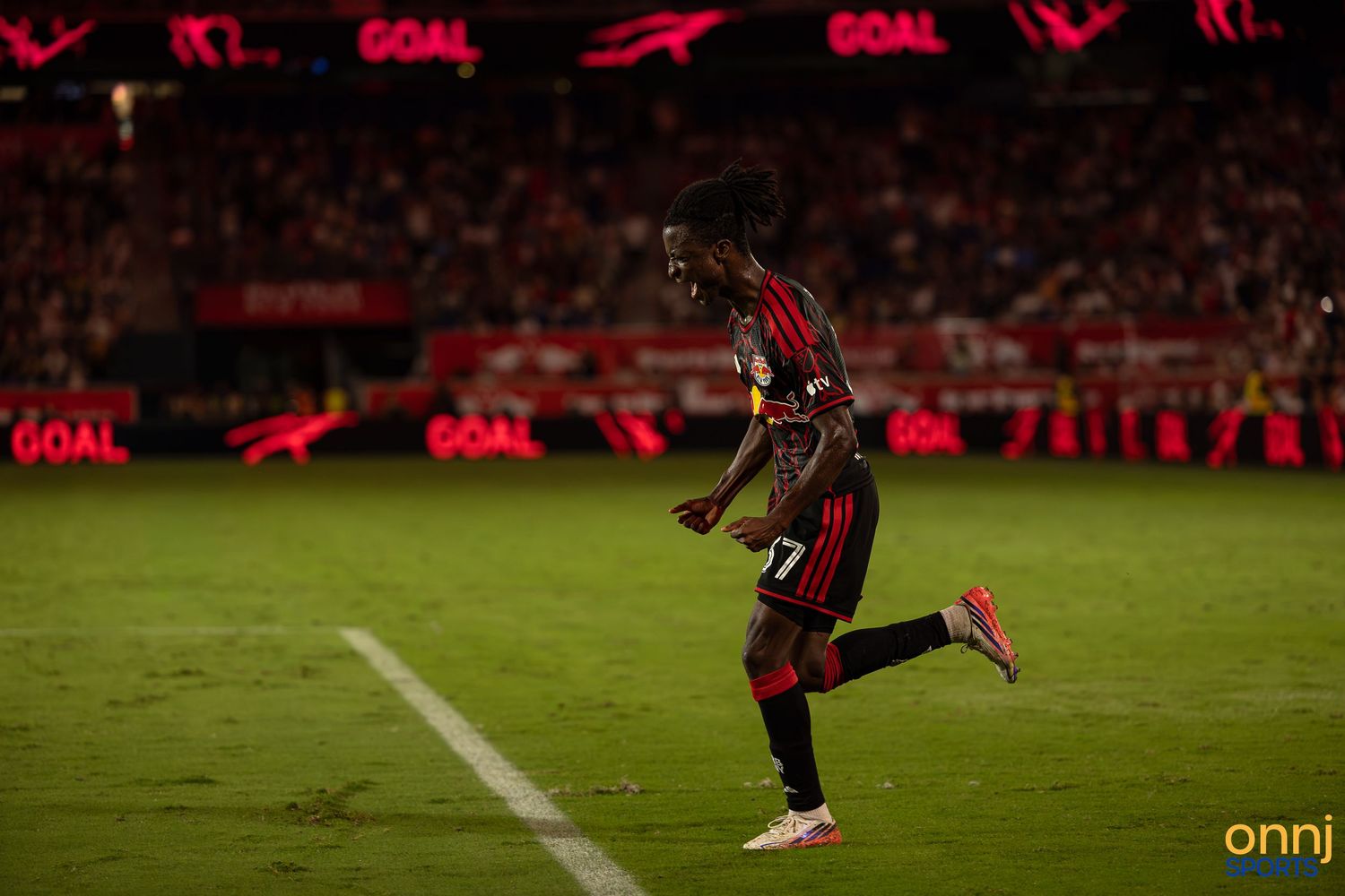 Soccer player celebrates a goal on the field at night, wearing a red and black jersey, with GOAL signs lit up behind.