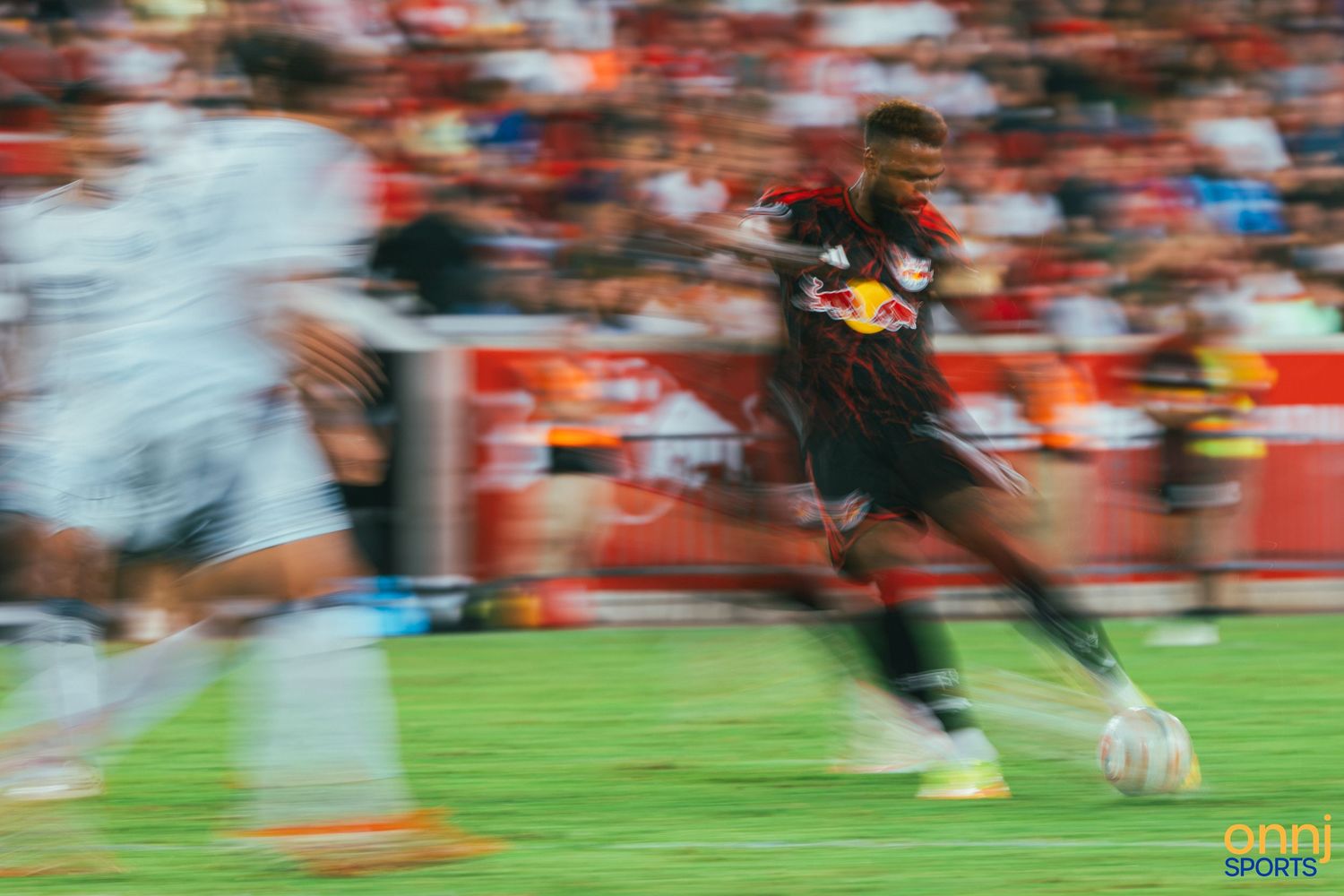 Soccer player in Red Bulls kit dribbles at speed during a fast-paced MLS match under stadium lights.