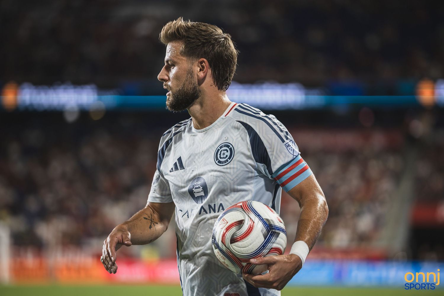 Soccer player in white jersey holds a match ball on the field during a professional MLS game at night.