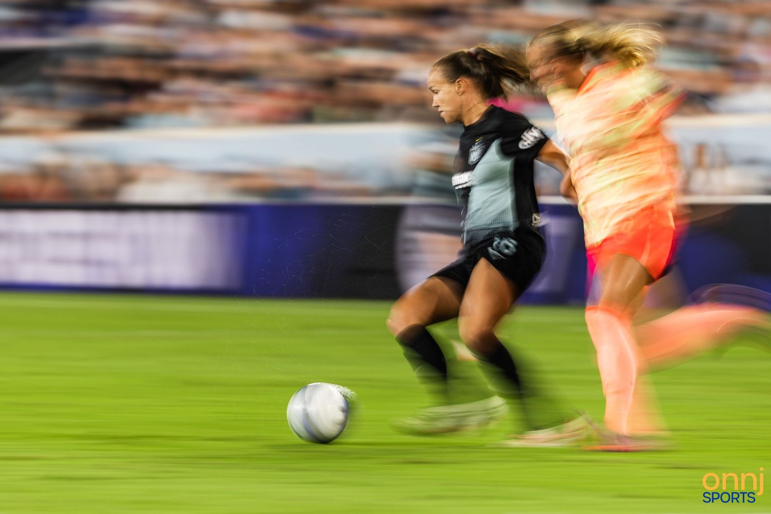 Two female soccer players compete for the ball during a night match, captured with motion blur effect.