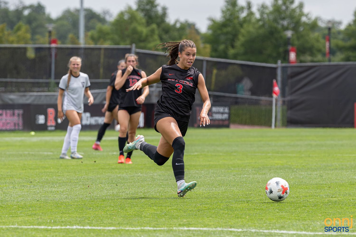 Female soccer player in red uniform chasing ball on green field during college soccer match.