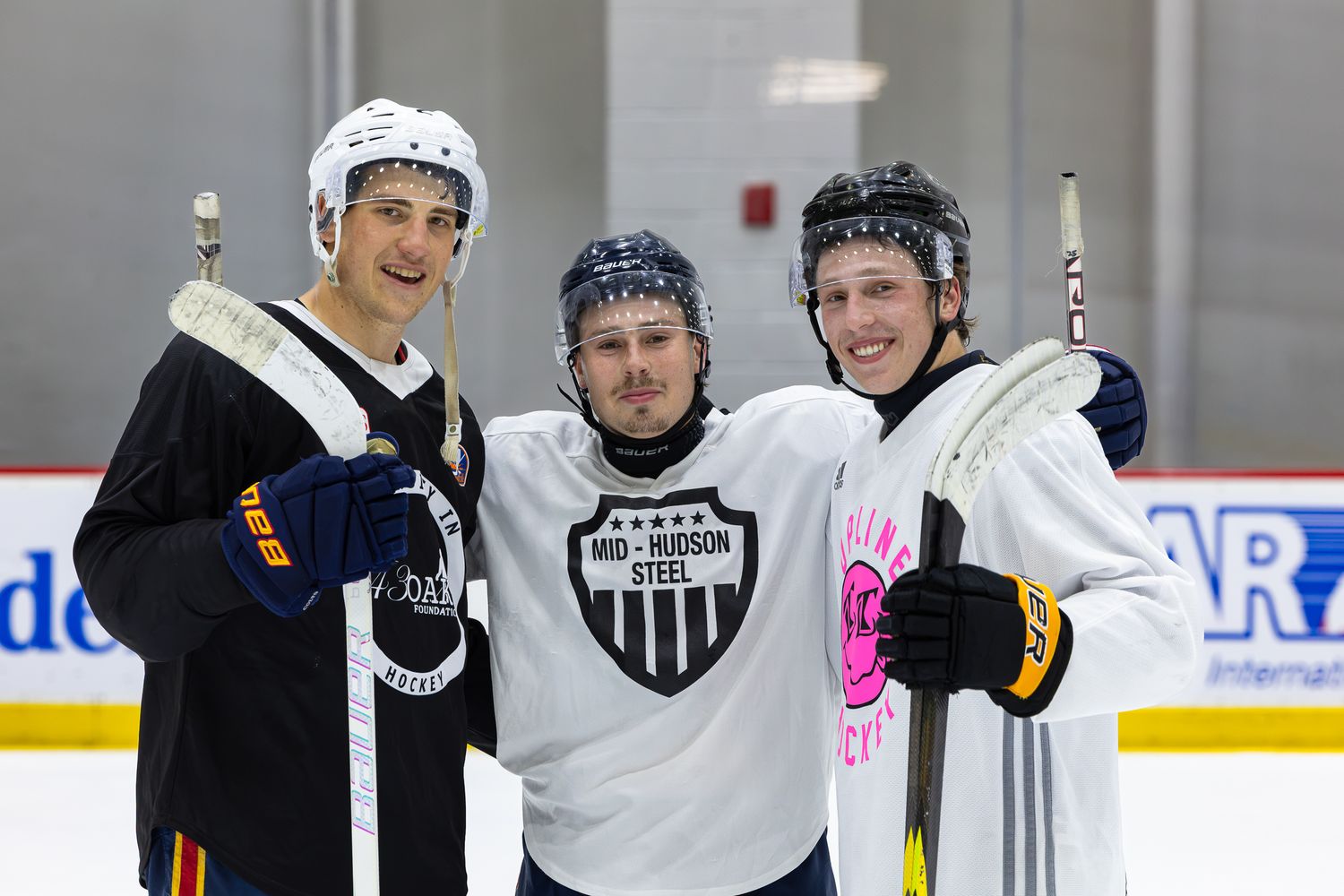 Three hockey players pose smiling on ice rink wearing Mid-Hudson Steel jerseys and holding sticks.