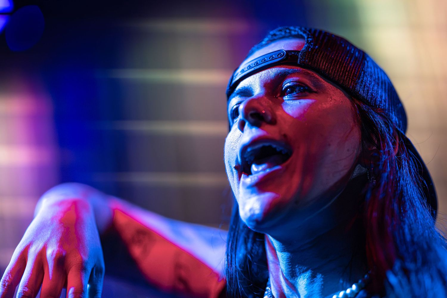 Rock performer with long hair and backwards cap singing passionately under dramatic blue and purple stage lighting.