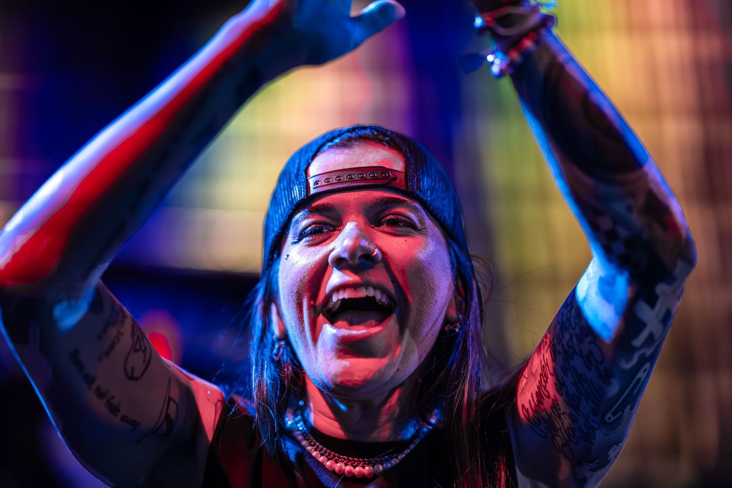 Tattooed rock performer with backward cap singing passionately on stage under dramatic red and blue lighting.