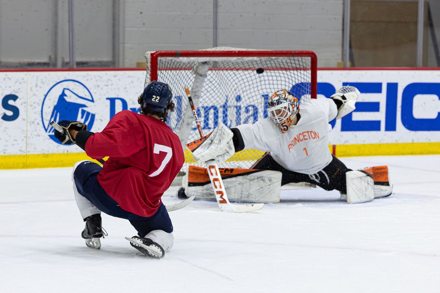 Hockey player in red jersey shoots on goalie in white during an indoor ice rink practice session.
