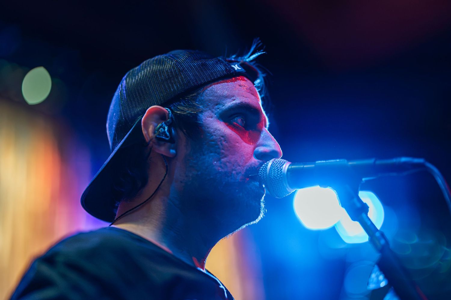 Male vocalist performing live on stage under blue and red lights, singing into a microphone wearing a backwards cap.