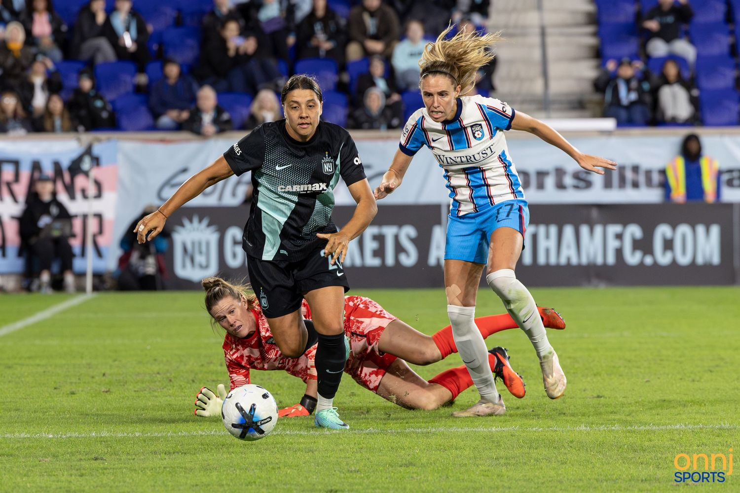 Women's soccer players compete in an intense match, with one player sliding and two others sprinting on a blue-bordered field.