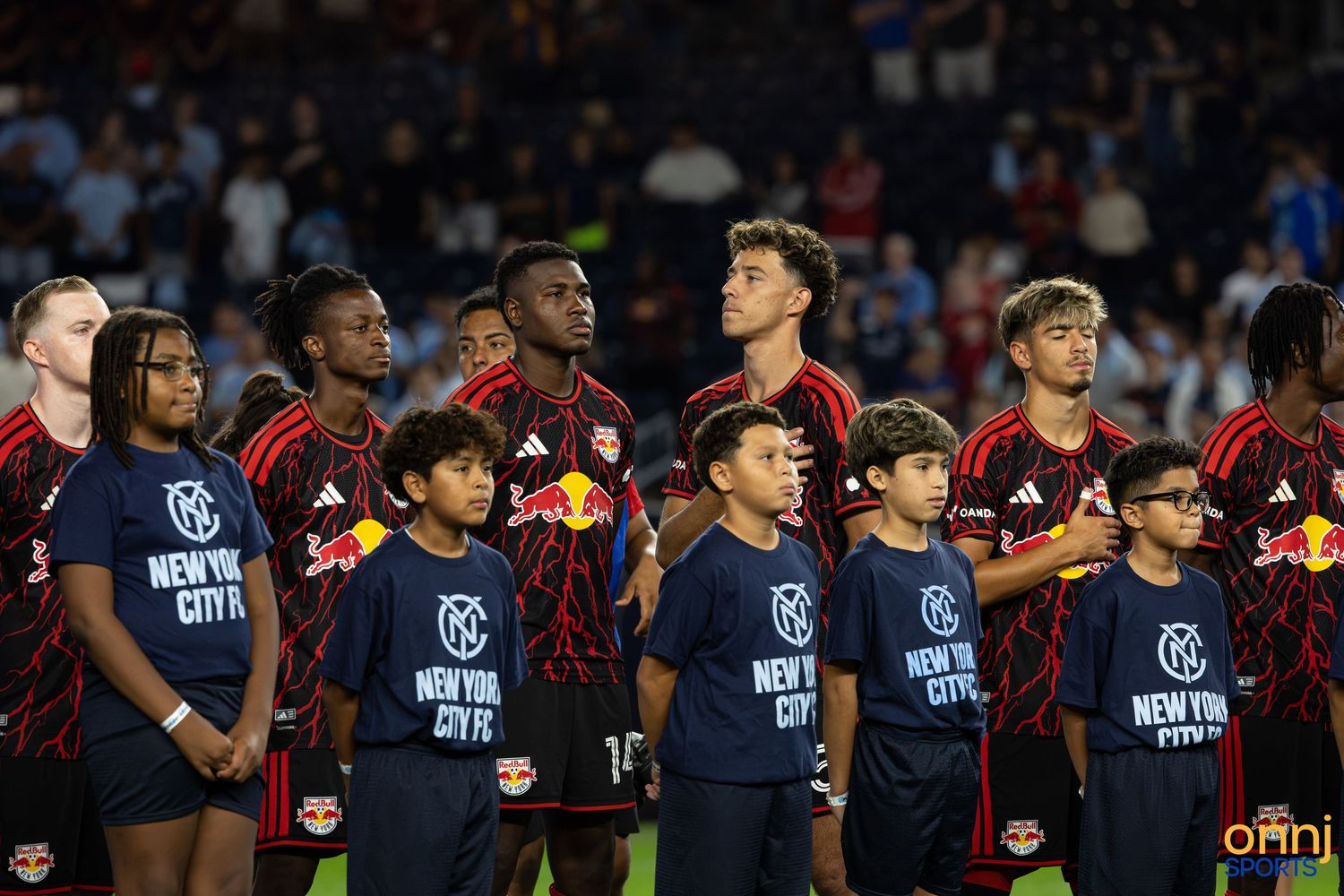 New York Red Bulls players line up with NYCFC youth escorts before an MLS soccer match at night.