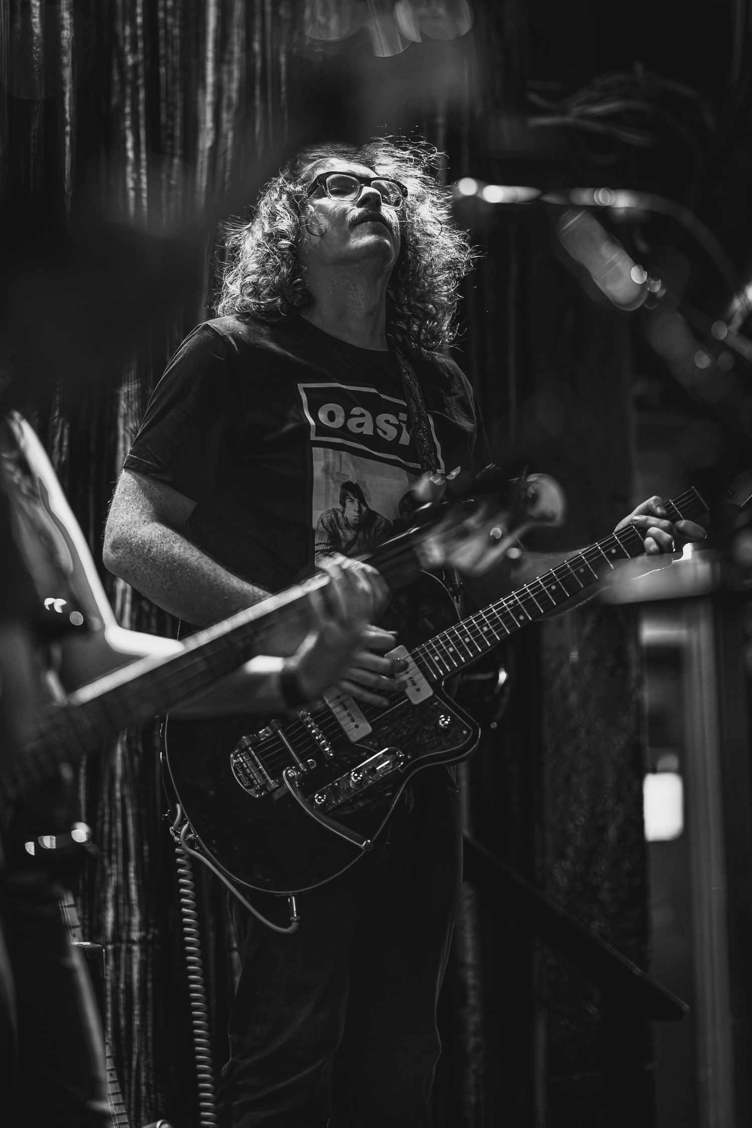 Black and white photo of a curly-haired guitarist in an Oasis t-shirt playing electric guitar on stage.