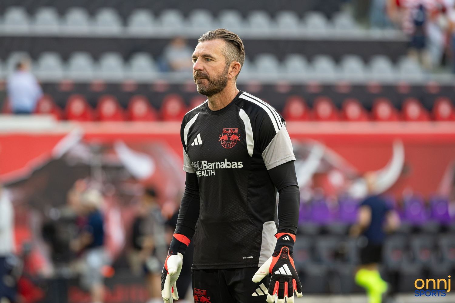 Soccer goalkeeper in red uniform with St. Barnabas Health sponsor stands on field during warm-ups.