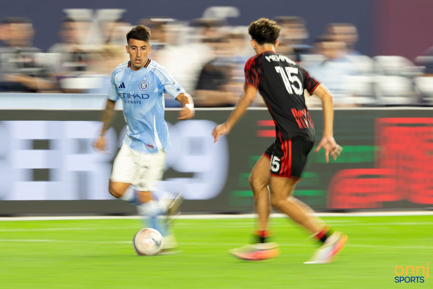 Two soccer players compete for the ball during a night match, one in light blue and one in black and red uniforms.