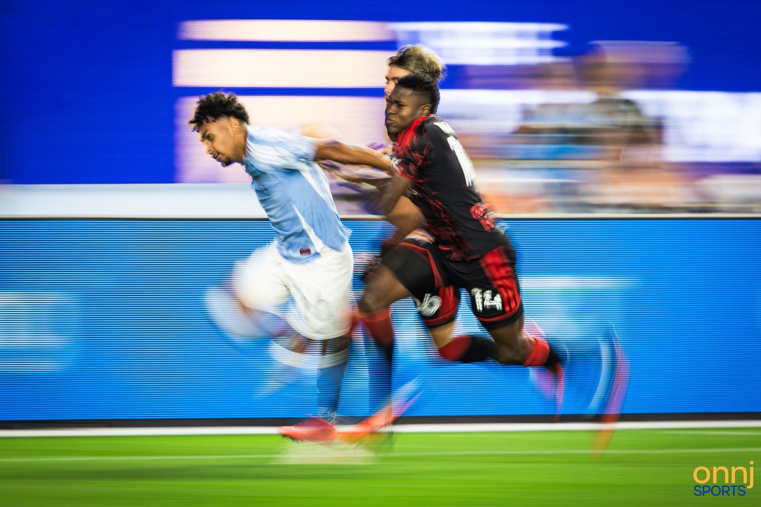 Two soccer players sprint in a dynamic blur during an intense match, one in light blue and one in red and black.