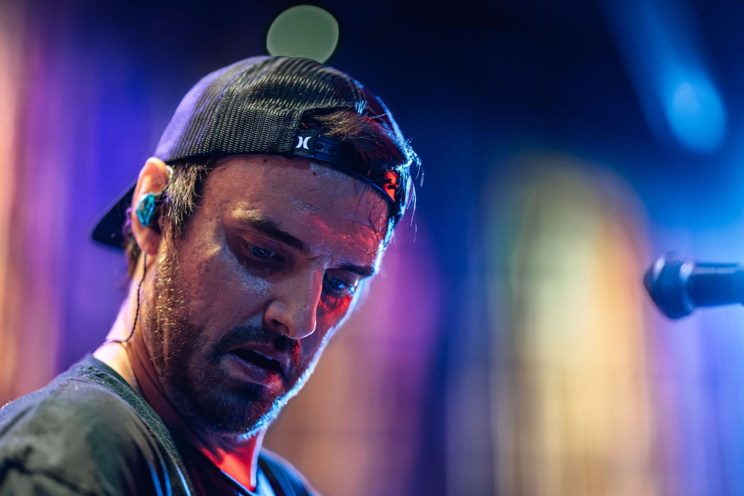 Male musician performing on stage wearing a backward cap with colorful stage lights in background.