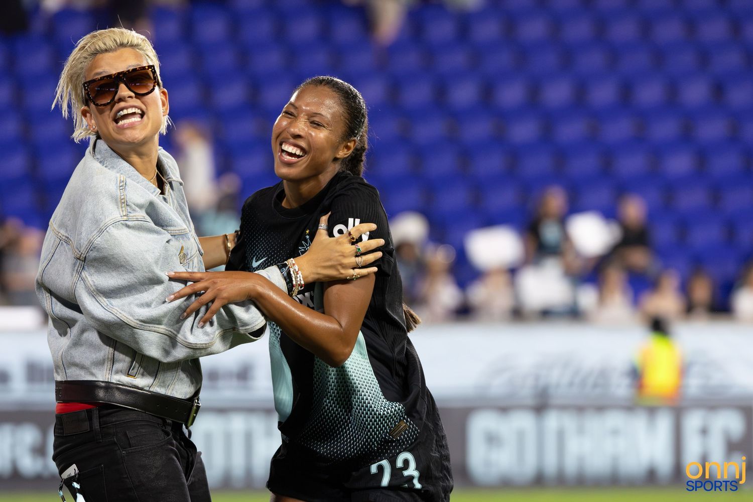 Two women laughing and embracing on a soccer field at a Gotham FC event during an outdoor game.