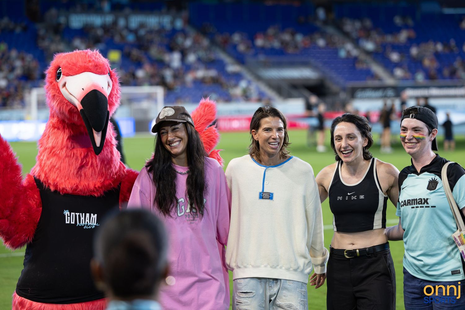 Gotham FC mascot poses with four smiling fans on the soccer field during a match at Red Bull Arena.