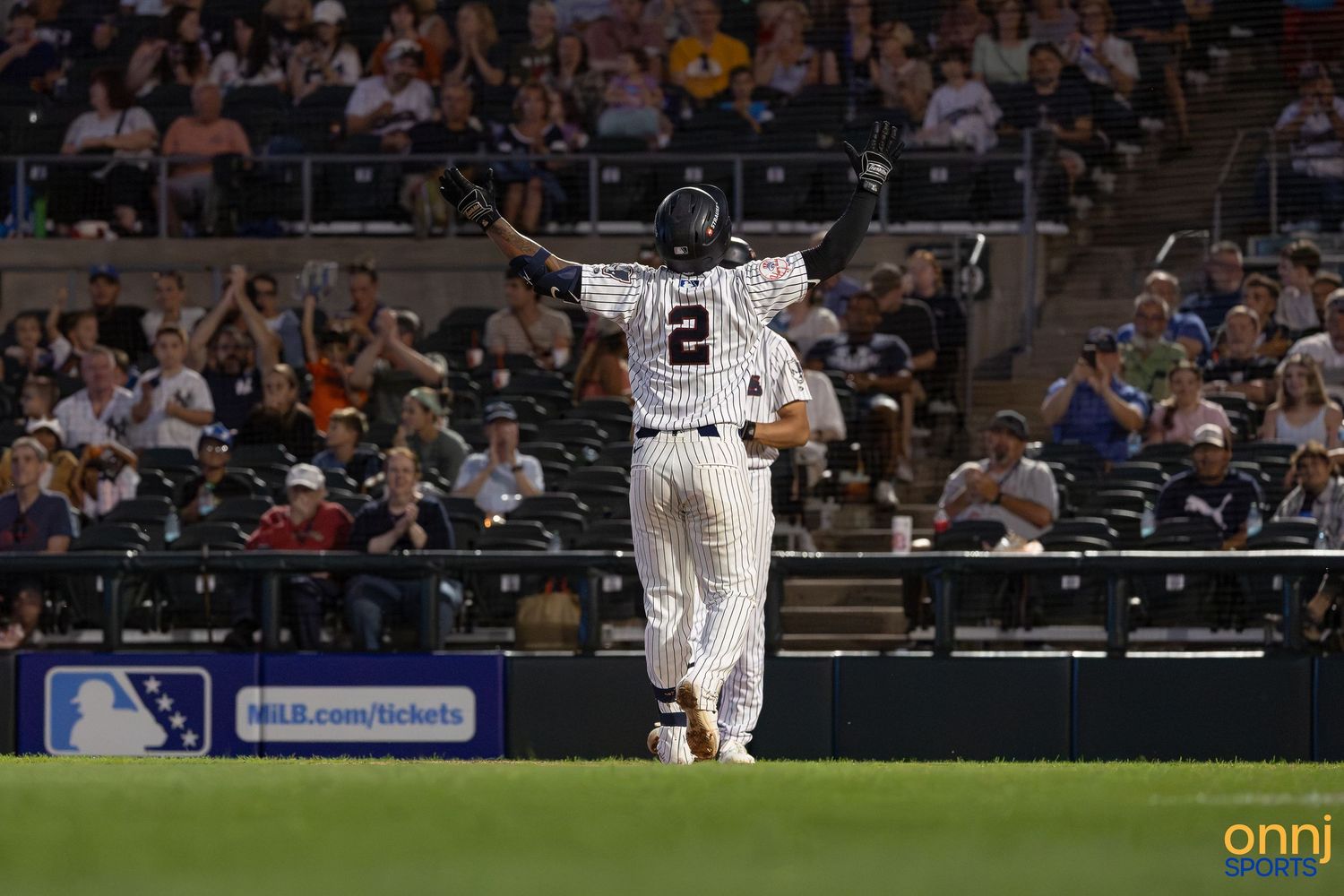 Baseball player #2 celebrates in pinstripe uniform at home plate, arms raised, with crowd cheering in stadium background.