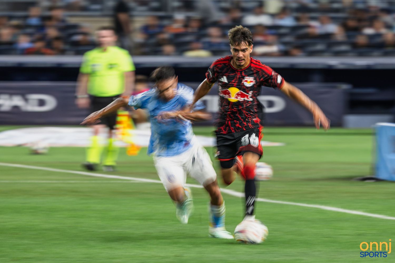 Two MLS soccer players compete for the ball during a New York Red Bulls vs NYFC match under stadium lights.