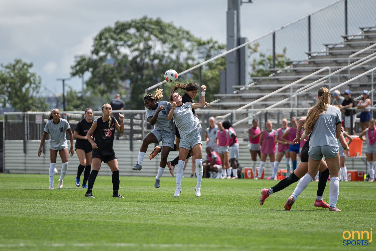 Women's soccer players compete for a header during an outdoor match on a green field with spectators in the background.