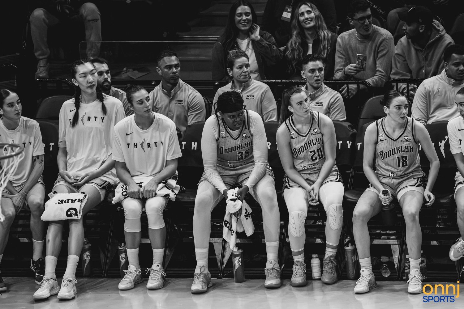 Black and white photo of a women's basketball team sitting on the bench during a game.