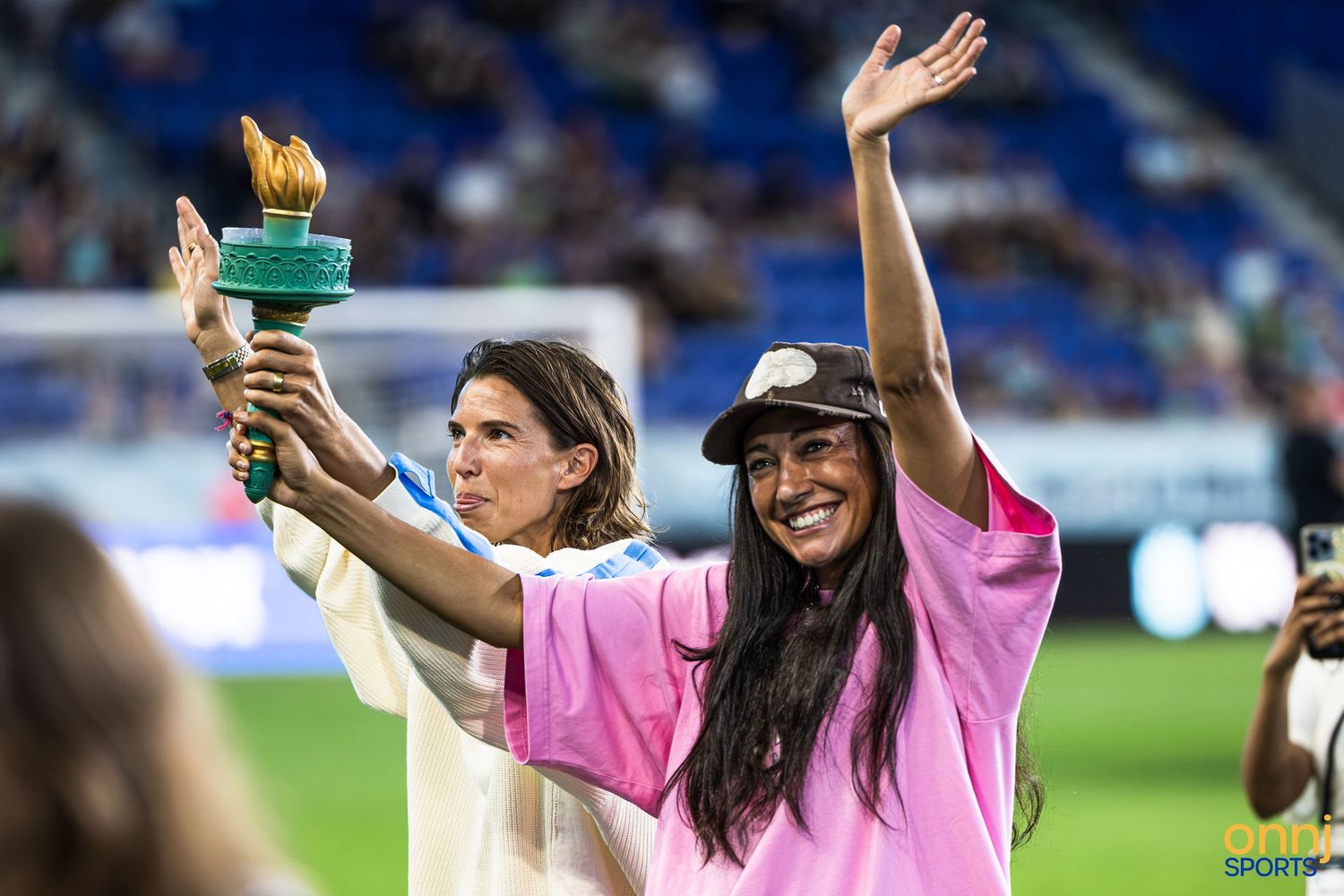 Two women celebrate on a soccer field, one holding a torch trophy, both waving to the crowd in a stadium.
