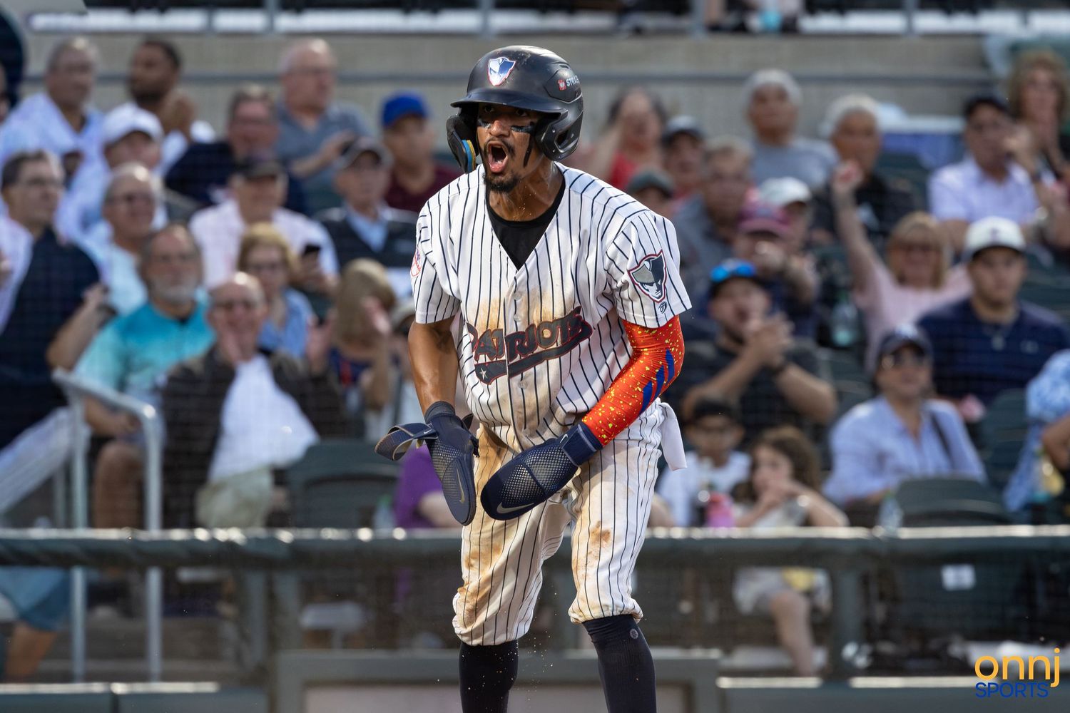 Baseball player in pinstripe uniform reacts after a hit during a sunny minor league game with fans in the background.