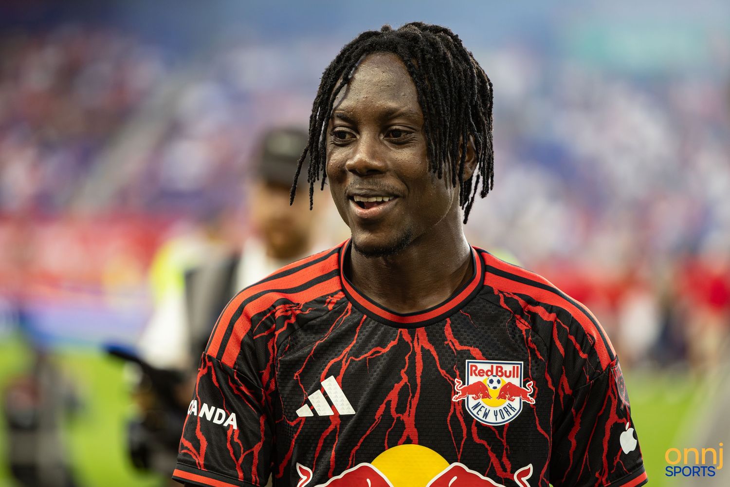 Young soccer player in Red Bulls black and red jersey smiling on field after MLS match.