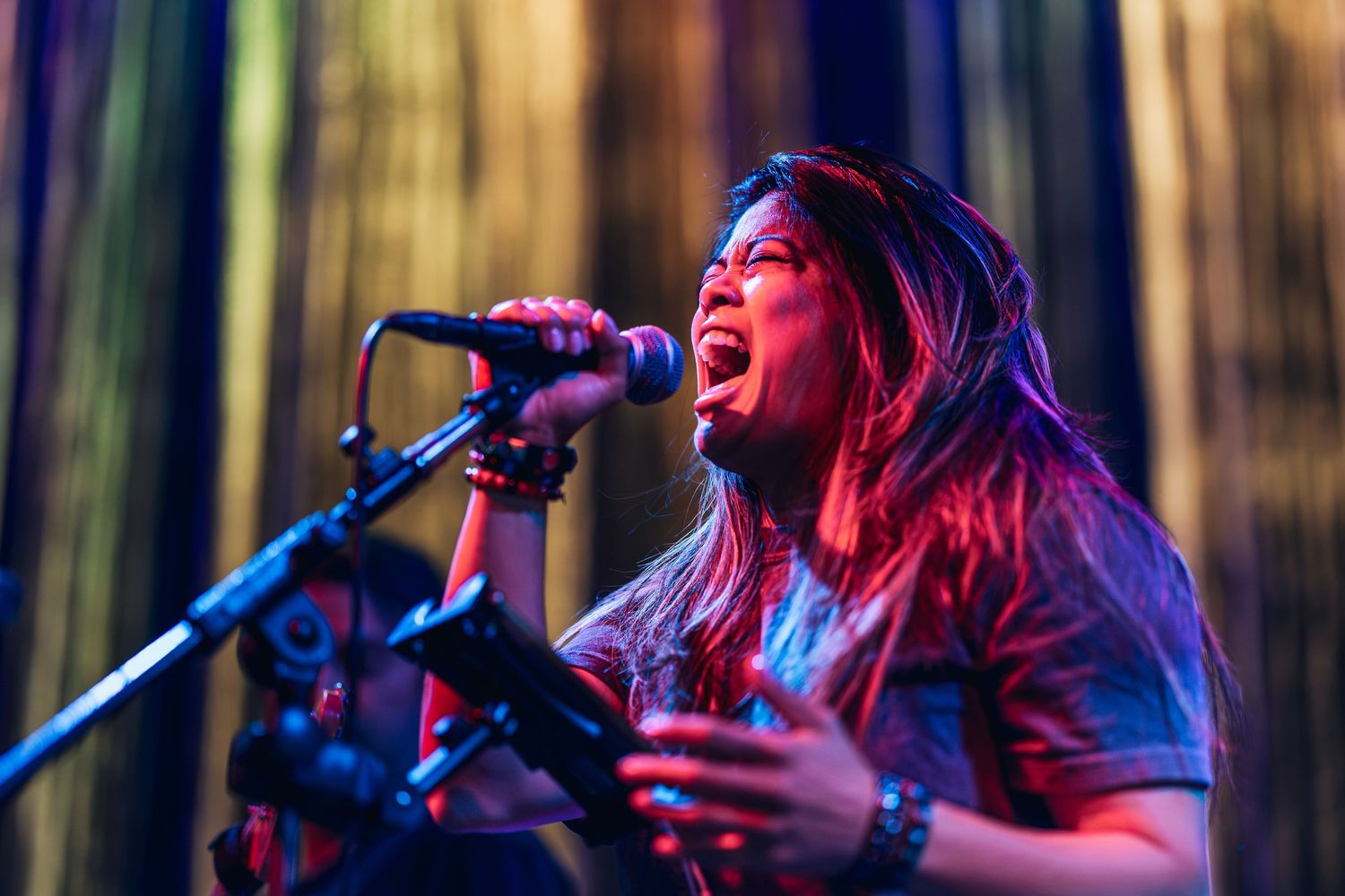 Female vocalist singing passionately into microphone on stage under vibrant red and blue concert lighting.