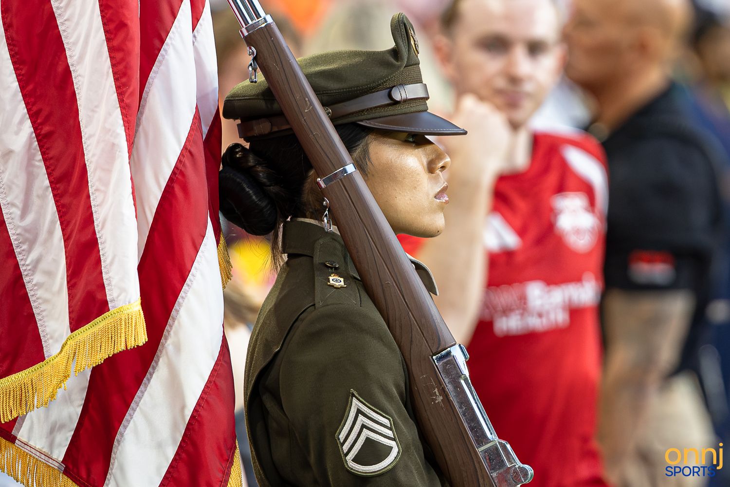 Female military soldier in dress uniform holding a rifle and American flag during a color guard ceremony at a sporting event.
