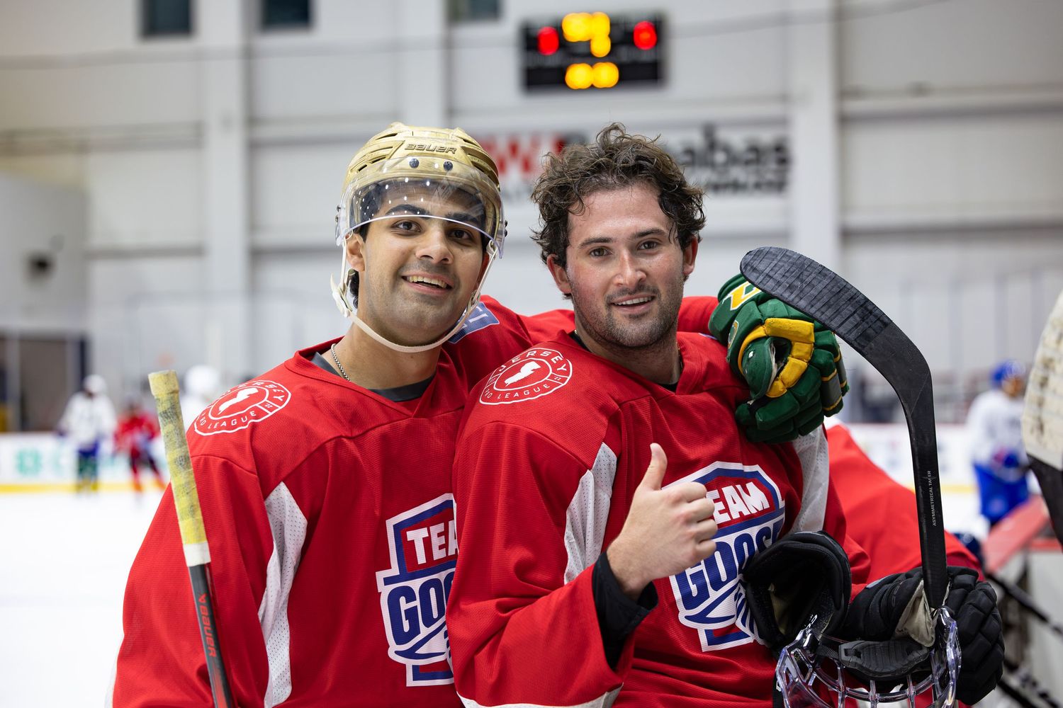 Two hockey players in red Team Goal jerseys smile together on the ice rink during a game.