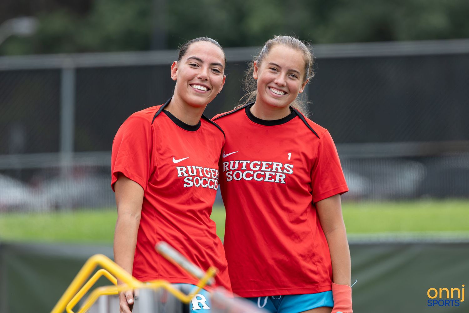 Two smiling Rutgers Soccer players in red jerseys pose together on an outdoor field during practice.
