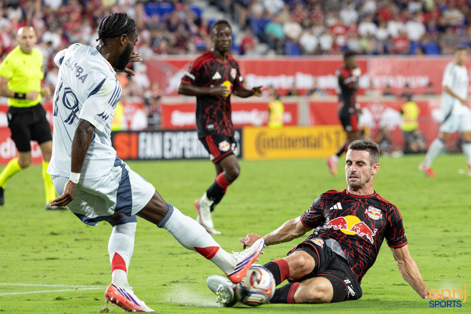 Two MLS soccer players compete for the ball during an intense match, one in white and one in a Red Bulls uniform.