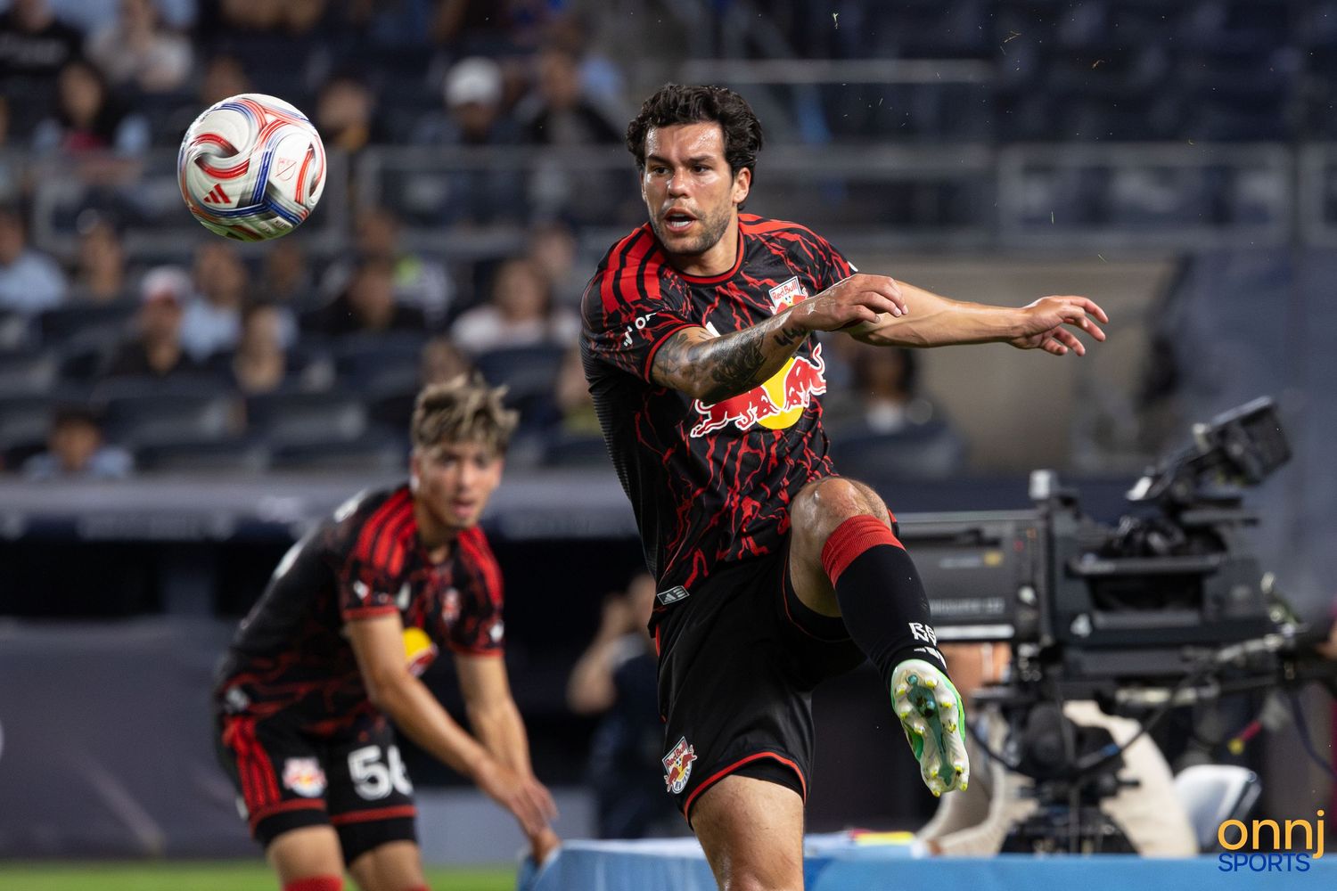 Soccer player in Red Bulls kit chases ball during a night match with teammates and camera crew visible.