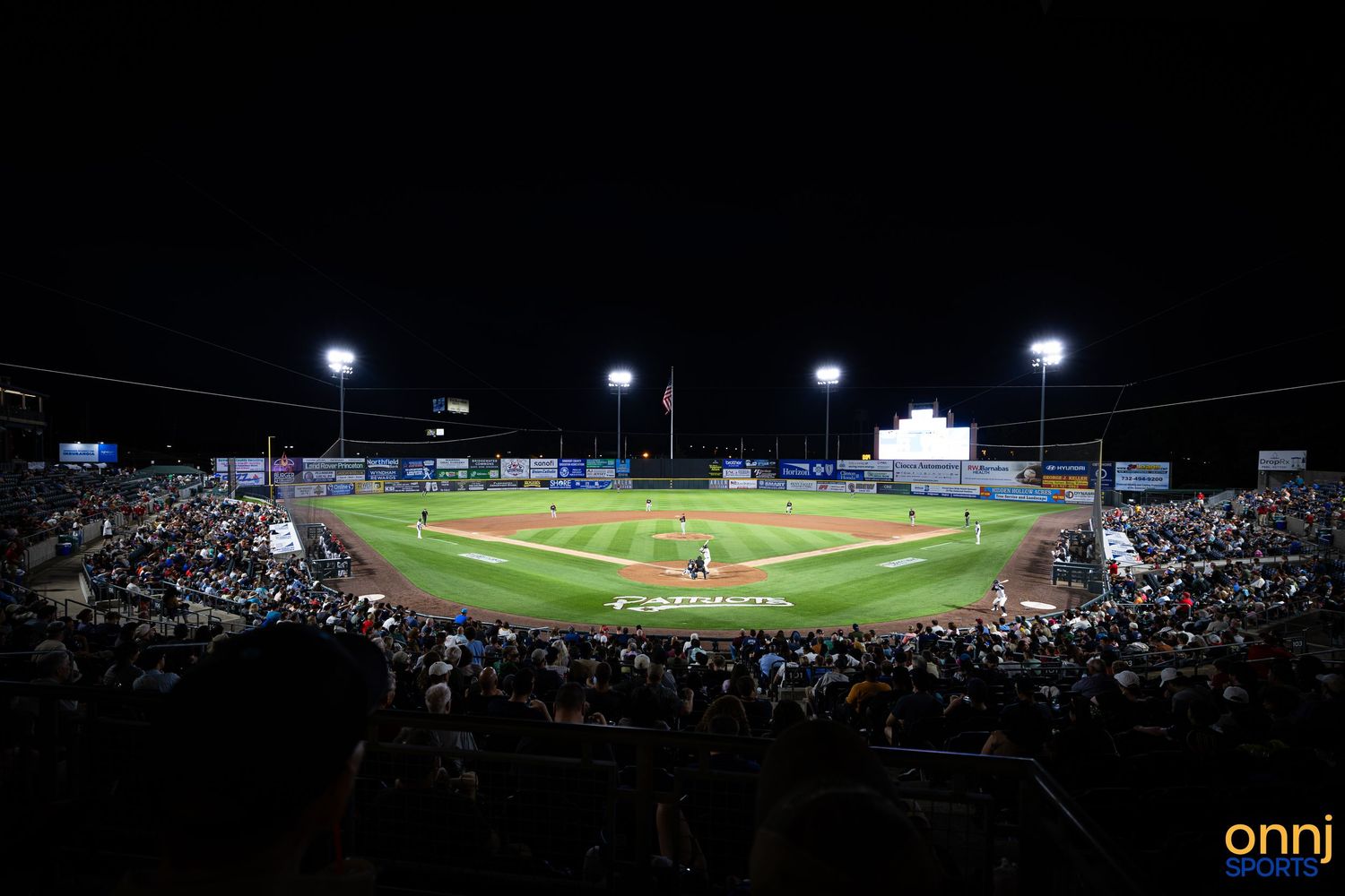 Night game at a minor league baseball stadium with bright lights illuminating the field and a large crowd in the stands.
