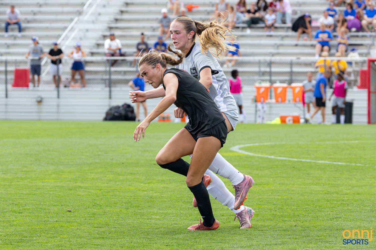Two female soccer players compete for the ball during a match on a green field with stadium seating in the background.