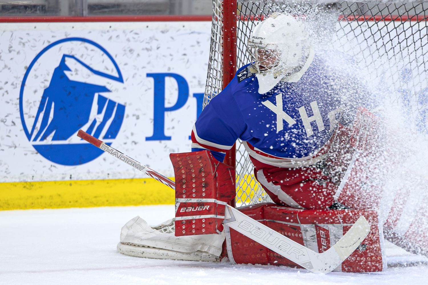 Hockey goalie in red and blue uniform makes a save at the net, sending ice flying during an intense game moment.