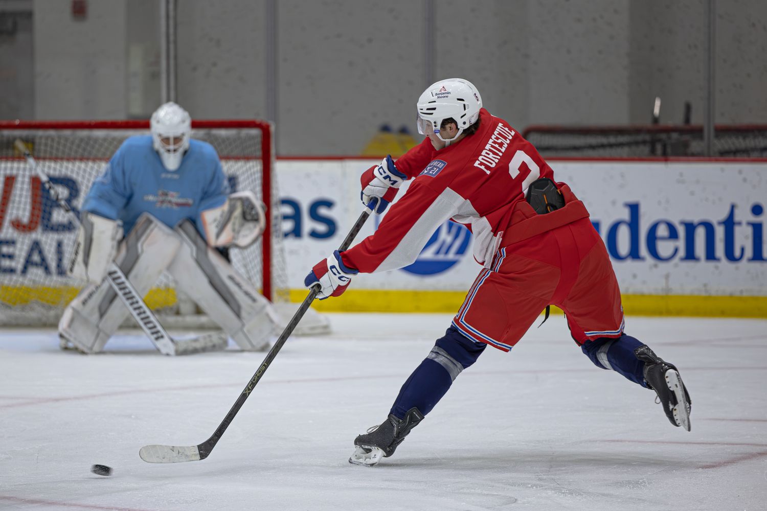 Hockey player in red jersey taking a shot on goal during an indoor ice rink practice session.
