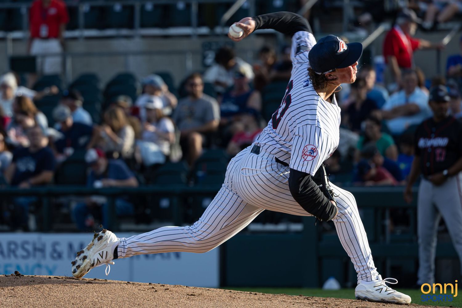 Baseball pitcher in white pinstripe uniform delivers a powerful pitch during a night game with fans in the background.