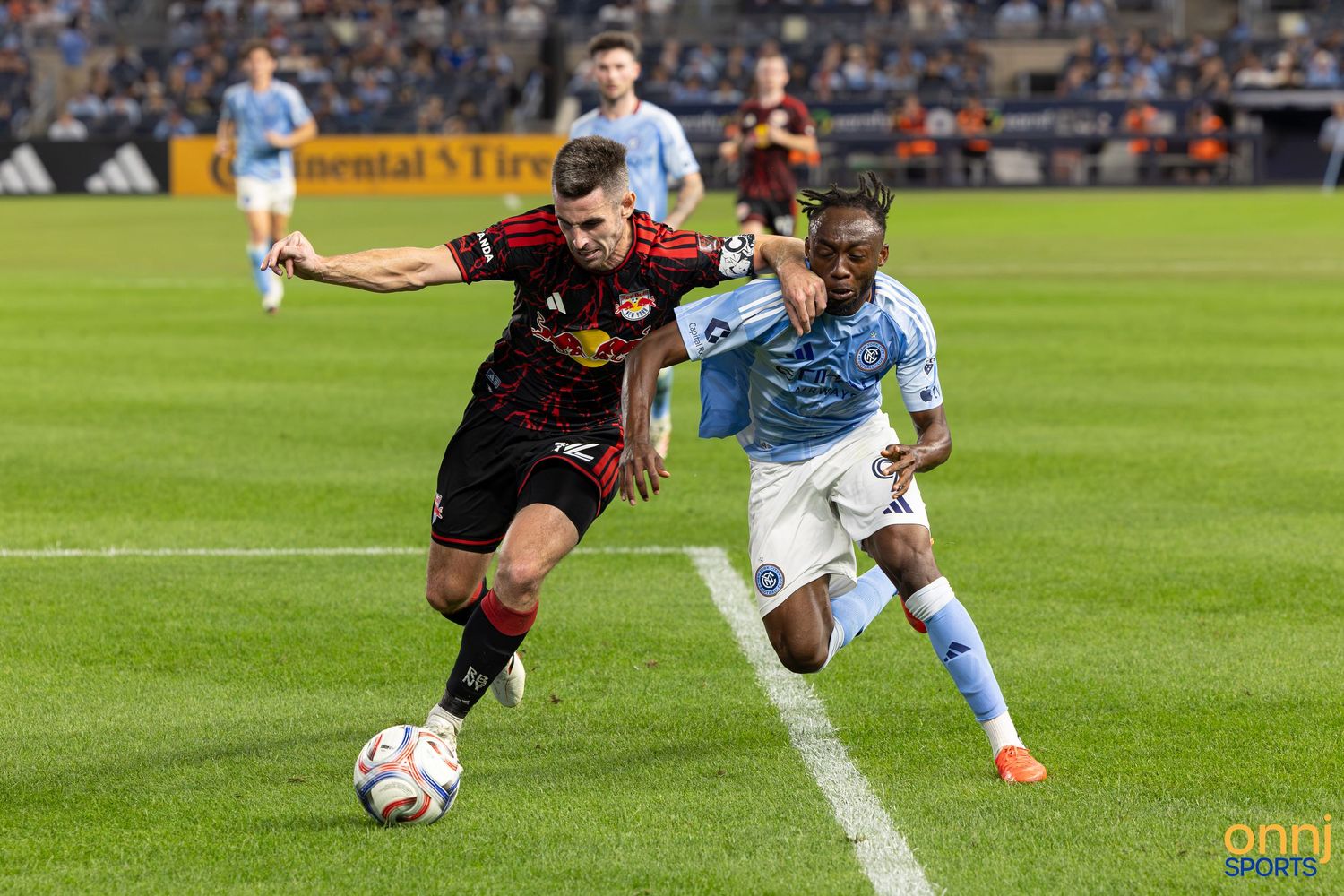 Two MLS soccer players battle for the ball during a competitive match between NY Red Bulls and NYCFC at night.