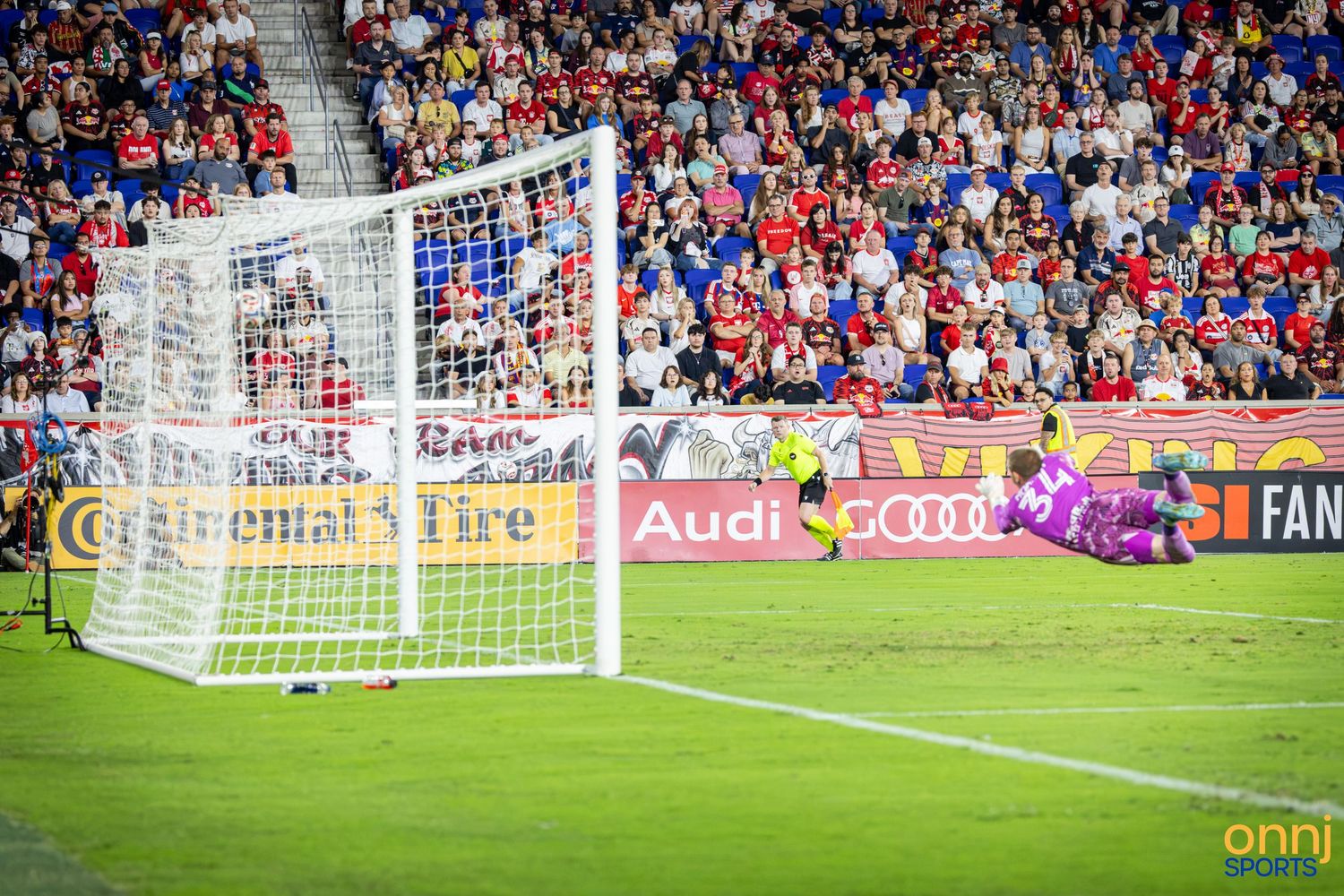 Soccer goalkeeper dives to make a save during an MLS match at Red Bull Arena with a packed crowd in the background.