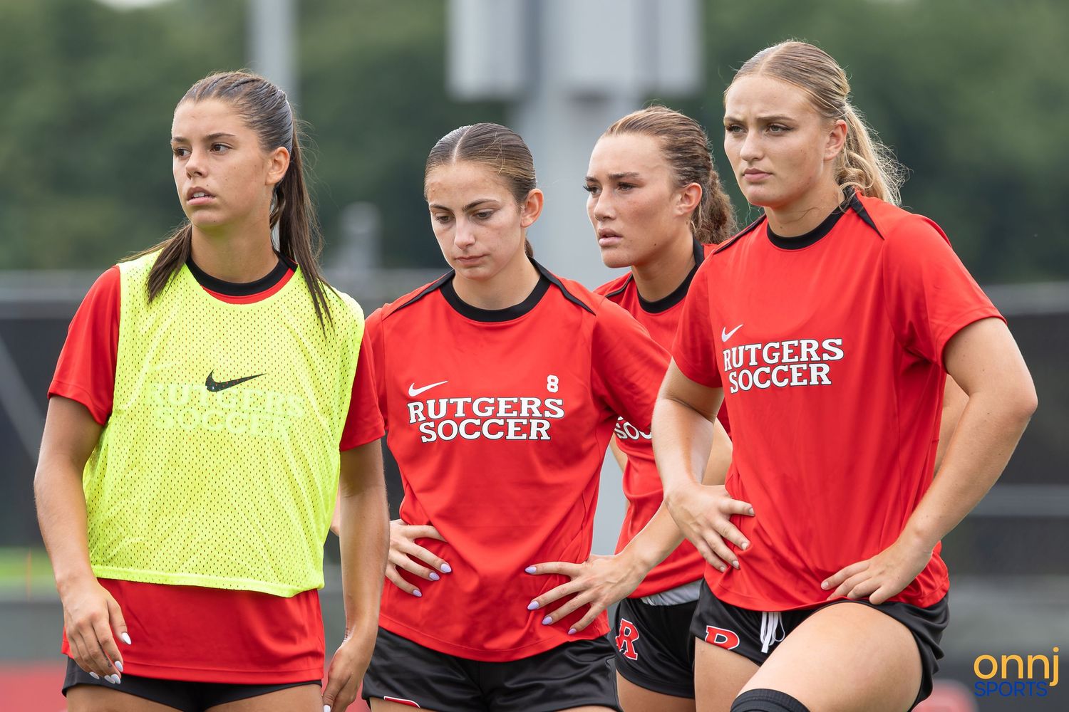 Four Rutgers Soccer female players in red uniforms stand together on a green field during practice.