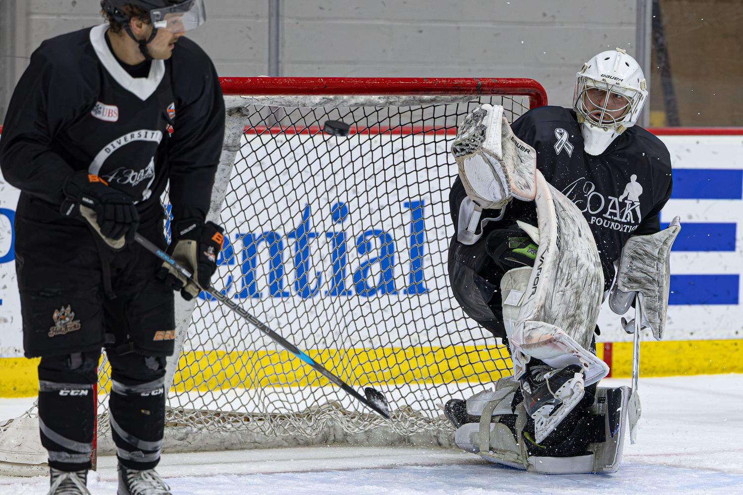 Two ice hockey players during practice, with a goalie in white gear defending the net on an indoor rink.