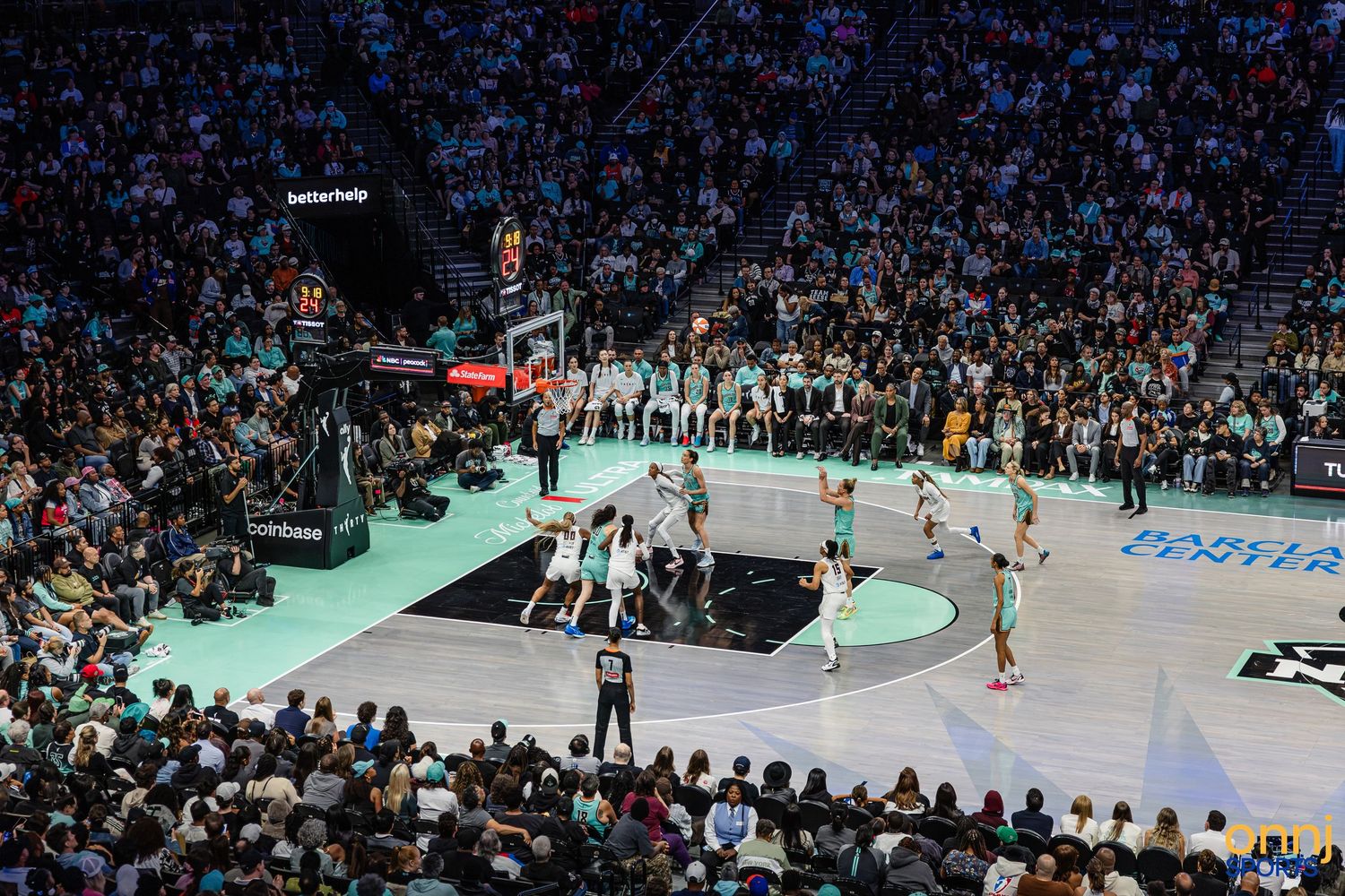 Packed arena crowd watches live WNBA basketball game on teal and black court under bright stadium lights.
