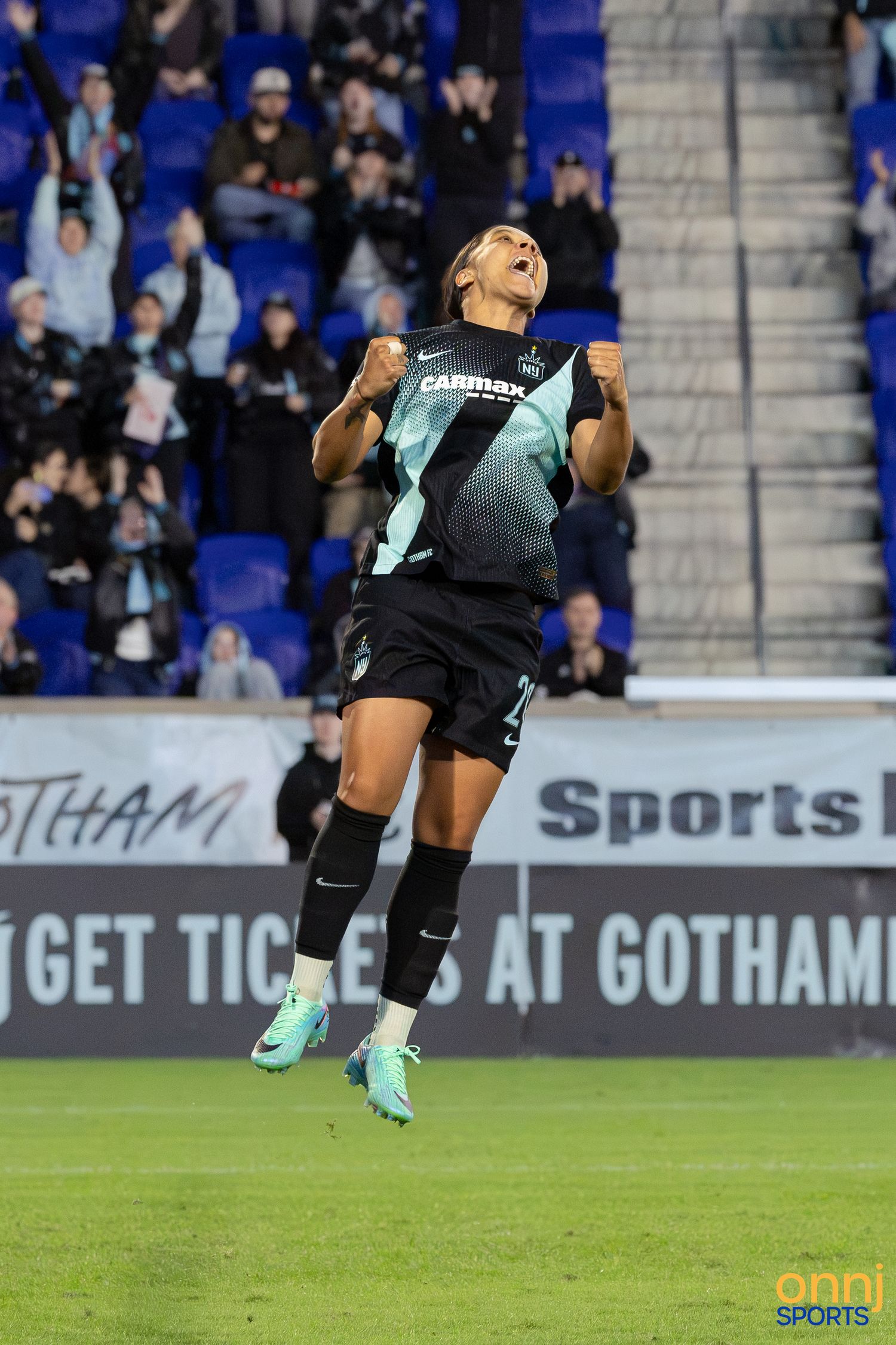 Soccer player in teal Gotham FC jersey leaps celebrating a goal at Red Bull Arena during an NWSL match.